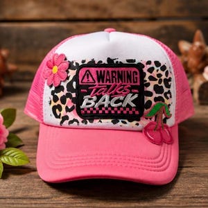May include: A pink and white trucker hat featuring a black rectangular patch with the text WARNING talks BACK. The front panel displays a black and white leopard print pattern, accented by a pink flower patch on the left and a cherry patch on the right.