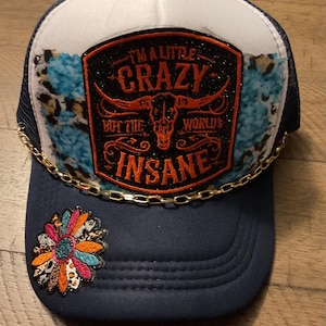 Può includere: Un cappellino trucker blu navy e bianco con una toppa centrale raffigurante un teschio di toro e il testo I'M A LITTLE CRAZY BUT THE WORLD'S INSANE. Il cappellino presenta una catena color oro sulla visiera e una toppa floreale multicolore.