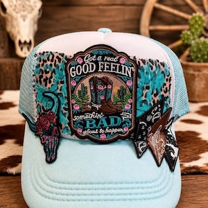 Peut inclure: Une casquette trucker bleu clair avec un écusson central brodé portant le texte Got a real GOOD FEELIN somethin BAD about to happen, illustrant des bottes de cow-boy et des cactus. Elle comporte des écussons latéraux en forme de crâne floral.