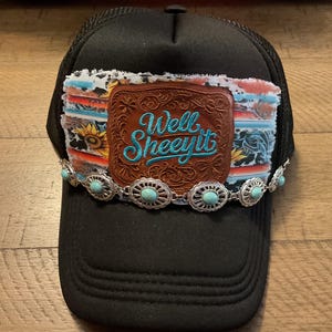 May include: A black trucker hat featuring a rectangular patch with a frayed edge, a sunflower and cow print pattern, and a brown leather center piece embossed with the text Well Sheeyit in blue. A silver-toned chain with turquoise stones accents the front.