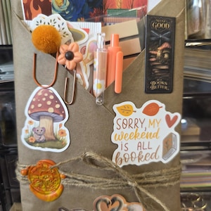 May include: A brown paper envelope filled with stationery items including orange pens, a pom-pom paperclip, a flower-shaped clip, and various stickers. A sticker reads, "SORRY, MY weekend is ALL booked" and a bookmark says, "SLEEP GOOD BUT BOOKS BETTER."