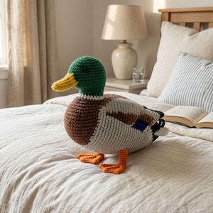 May include: A crocheted mallard duck plush toy featuring a vibrant green head, a bright yellow beak, a brown chest, and a light gray body with black and blue wing details. The duck has orange webbed feet and a small black button eye.