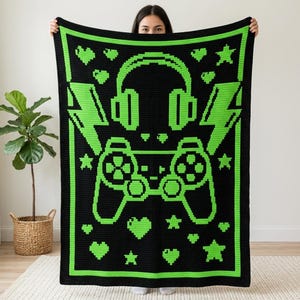 Pode incluir: Uma manta de crochê retangular com fundo preto e um design pixelado em verde neon vibrante. O padrão apresenta um controle de videogame central, fones de ouvido, raios, estrelas e formas de coração.
