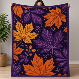 May include: A rectangular crochet blanket featuring a dark purple background with a repeating pattern of large maple leaves in vibrant orange, yellow, and purple hues. The blanket has a solid dark purple border and a textured, woven appearance.