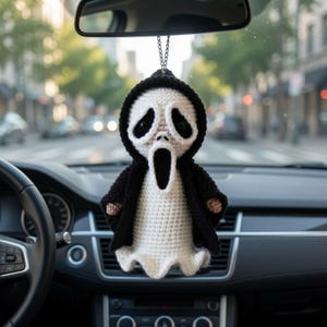 May include: A crocheted hanging ornament featuring a ghost-like figure with a white face, large black eye sockets, and an open black mouth, wearing a black hooded cloak. The figure is suspended by a silver-toned metal chain from a rearview mirror.