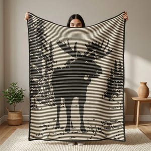 May include: A rectangular knit throw blanket featuring a black and cream horizontal striped pattern. The design displays a large silhouette of a moose standing in a forest setting, created by the contrast between the black and cream yarn colours.