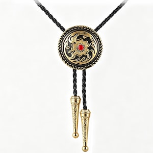 Könnte beinhalten: Eine Bolo-Krawatte mit einem runden, gold-schwarzen Medaillon, das ein graviertes Blumenmuster und einen zentralen roten Stein aufweist. Die schwarze geflochtene Kordel endet in zwei passenden goldfarbenen Metallspitzen mit aufwendiger Gravur.