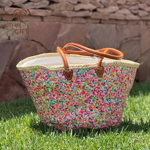 Pode incluir: Uma bolsa tipo cesta de palha trançada coberta com pequenas lantejoulas multicoloridas em tons de rosa, verde, azul e amarelo. A bolsa possui duas alças de couro castanho claro presas com abas de couro costuradas e um forro de tecido branco.