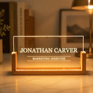 May include: A rectangular clear acrylic desk sign with a light-colored wooden base. The sign is illuminated from the bottom and features the white text JONATHAN CARVER and MARKETING DIRECTOR with horizontal lines above and below the job title.
