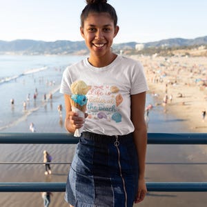 Puede incluir: Una camiseta blanca de manga corta con un estampado gráfico de un cono de helado y varias conchas marinas con el texto Life is better at the beach, combinada con una minifalda de mezclilla azul oscuro con una cremallera frontal vertical.
