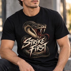 Peut inclure: Un t-shirt noir à manches courtes orné d'un motif représentant un serpent enroulé et agressif, la gueule ouverte, les crocs exposés et la langue rouge. Sous le serpent, le texte STRIKE FIRST est imprimé dans une police blanche et usée.