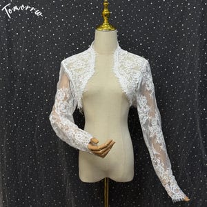 May include: A white lace bolero jacket featuring long sleeves and intricate floral embroidery patterns. The garment has an open front design with scalloped edges along the neckline and cuffs, displayed on a mannequin. The text Tomorrow is visible.