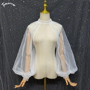 May include: A white sheer tulle bolero featuring a high neckline and voluminous bishop sleeves that gather at the wrists. The garment is displayed on a mannequin, showcasing the translucent fabric and the structured, airy silhouette of the sleeves.