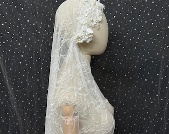 May include: A white lace bridal veil featuring intricate floral embroidery and small pearl accents. The veil is draped over a mannequin head and shoulders, showcasing a delicate scalloped edge and layered fabric construction with floral applique.