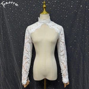 May include: A white lace bolero with long sleeves, a high mock neck collar, and a cutout chest design. The sleeves feature small white buttons at the cuffs. The garment is displayed on a beige mannequin. The text Tomorru is visible in the top left.
