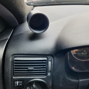Puede incluir: Un soporte cilíndrico para manómetro en color negro mate colocado sobre el salpicadero texturizado de un vehículo, situado directamente encima de la rejilla de ventilación horizontal y el interruptor de luces en el lado del conductor.