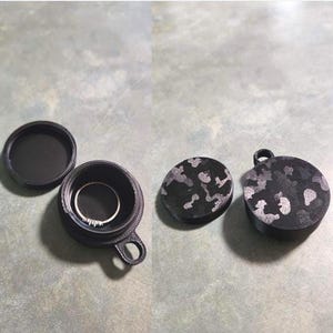May include: A small, round, black 3D-printed container with a lid, featuring a camouflage pattern on the exterior. The container has a small loop for attachment and holds a thin silver ring with small stones inside the open compartment.