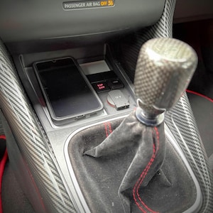 Puede incluir: Una consola central de coche con pomo de palanca de cambios de fibra de carbono, funda de ante negro con costuras rojas y un smartphone en un compartimento. La consola incluye una toma de 12V, un puerto USB y un botón ESP OFF.