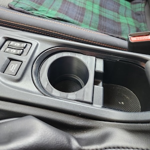May include: A black 3D-printed cup holder insert designed for a vehicle center console. The circular insert features a textured surface and fits into the existing console space next to a button labeled X-MODE and seat heater controls.