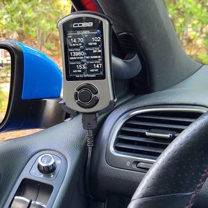 Puede incluir: Un dispositivo de diagnóstico automotriz COBB Accessport de color plateado y negro montado en el tablero de un automóvil. La pantalla muestra datos del motor en tiempo real, incluyendo AFR, voltaje de batería y temperatura del refrigerante.