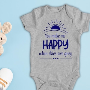 Peut inclure: Un body gris à manches courtes pour bébé avec un motif bleu foncé représentant un soleil et le texte "You make me HAPPY when skies are grey". Sont inclus un anneau en perles de bois, un hochet lapin en crochet et deux chaussures blanches.