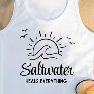 May include: A white tank top featuring a black graphic of a sun rising over a wave with birds flying nearby. Below the graphic, the text reads Saltwater in a cursive script font, followed by the words HEALS EVERYTHING in a bold sans-serif font.