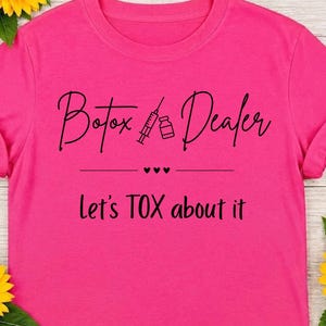 Può includere: Una t-shirt rosa acceso a maniche corte con la scritta nera Botox Dealer accompagnata da un'icona di siringa e flacone, seguita da tre piccoli cuori e dal testo Let's TOX about it stampato in un semplice carattere nero senza grazie.