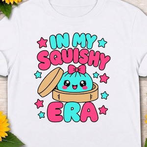 Könnte beinhalten: Ein weißes T-Shirt mit einer Grafik eines lächelnden, türkisfarbenen Knödel-Charakters mit einer rosa Schleife, der in einem Bambusdämpfer sitzt. Der Text "IN MY SQUISHY ERA" ist in fetten rosa und türkisfarbenen Buchstaben gedruckt, umgeben von kleinen Sternen.