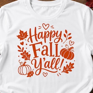 May include: A white short-sleeved t-shirt featuring a central orange graphic with the text Happy Fall Y'all. The design includes illustrations of two pumpkins, several maple leaves, small hearts, and sprigs, all rendered in a bold orange color.