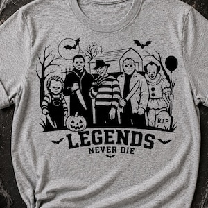 Könnte beinhalten: Ein graues, meliertes Kurzarm-T-Shirt mit einem schwarzen Grafikdruck von fünf Horrorfilm-Charakteren vor einem Haus, Bäumen und einem Vollmond. Unter den Charakteren steht der Text LEGENDS NEVER DIE in fetten, schwarzen Buchstaben.