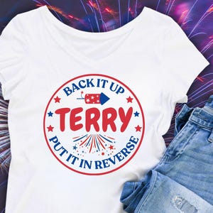 May include: A white V-neck t-shirt featuring a circular graphic with red and blue text that reads BACK IT UP TERRY PUT IT IN REVERSE, accompanied by a firework rocket icon, small stars, and a firework burst design in red and blue colors.