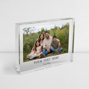 May include: A clear rectangular acrylic photo block featuring a printed photograph of a family of four sitting in a grassy field. The bottom section of the block includes the black printed text YOUR TEXT HERE and the smaller subtext Text Here.