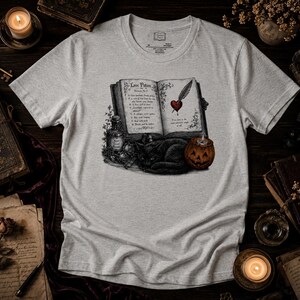 Può includere: Una t-shirt grigio chiaro a maniche corte con una stampa grafica di un libro aperto intitolato Love Potion, un gatto nero, una piccola bottiglia e una zucca intagliata. L'illustrazione del libro include un cuore trafitto da una penna.