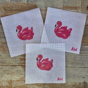 May include: Three square needlepoint canvases with a pink flamingo design. Each canvas has a white background and a flamingo in shades of pink. The initials "AW" are in the bottom right corner of two canvases. The canvases are on a wooden surface.