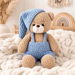 May include: A crocheted teddy bear with light brown yarn, featuring a cream-colored muzzle and a dark brown nose. The bear wears a blue crocheted nightcap with a white pom-pom and matching blue crocheted overalls with shoulder straps.