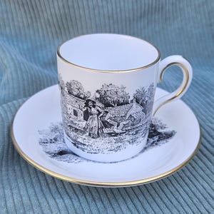 Pode incluir: Um conjunto de chávena e pires de porcelana branca com uma ilustração a preto e branco de uma pessoa num cenário rural com um ganso. A chávena e o pires têm um rebordo fino dourado nas extremidades e na pega.