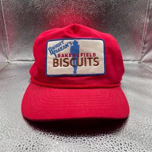 May include: A bright red baseball cap featuring a rectangular patch on the front. The patch has a cream background with a blue border and displays the text Dwight Yoakam's Bakersfield Biscuits in blue and brown, along with a blue silhouette figure.
