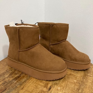 May include: A pair of ankle-height boots in a solid tan color with a suede-like exterior finish. These boots feature a thick, flat platform sole in a matching tan shade and a visible white faux-fur lining along the top edge of the boot opening.