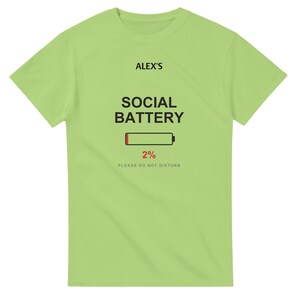 以下が含まれることがあります： 明るいライムグリーンの半袖Tシャツ。前面中央に黒字で「ALEX'S SOCIAL BATTERY」と書かれ、その下に赤色で2%と表示されたバッテリーアイコンと、小さなグレーの文字で「PLEASE DO NOT DISTURB」とプリントされています。