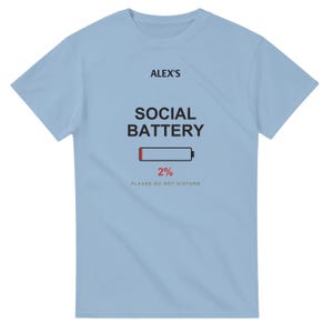 以下が含まれることがあります： ライトブルーの半袖Tシャツ。前面中央に黒文字で「ALEX'S SOCIAL BATTERY」、その下に赤いセグメント付きのバッテリーアイコン、赤い文字で「2%」、グレーの文字で「PLEASE DO NOT DISTURB」とプリントされています。