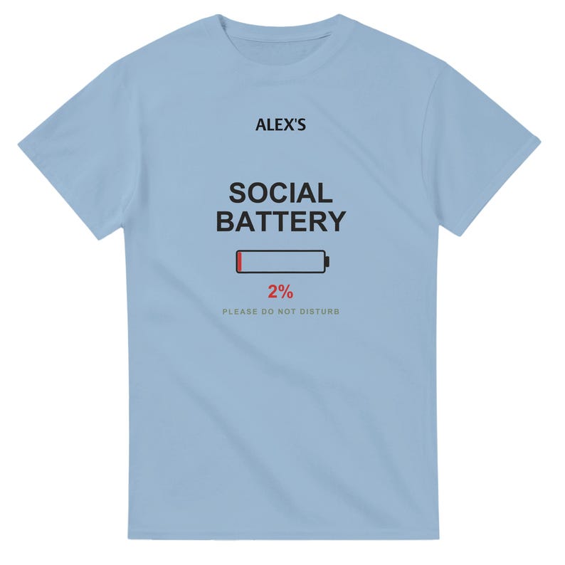 以下が含まれることがあります： ライトブルーの半袖Tシャツ。前面中央に黒文字で「ALEX'S SOCIAL BATTERY」、その下に赤いセグメント付きのバッテリーアイコン、赤い文字で「2%」、グレーの文字で「PLEASE DO NOT DISTURB」とプリントされています。