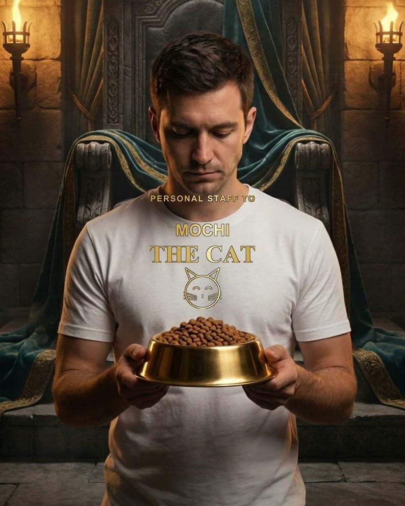 Puede incluir: Una camiseta blanca con el texto dorado PERSONAL STAFF TO MOCHI THE CAT y un icono de cara de gato dorado. La prenda es usada por una persona que sostiene un cuenco dorado lleno de comida seca marr&oacute;n. El dise&ntilde;o est&aacute; centrado en el pecho.