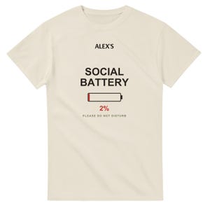 以下が含まれることがあります： クリーム色の半袖Tシャツ。前面中央に黒字で「ALEX'S SOCIAL BATTERY」、赤いバーが表示されたバッテリーアイコン、赤字で「2%」、黒字で「PLEASE DO NOT DISTURB」というテキストがプリントされています。