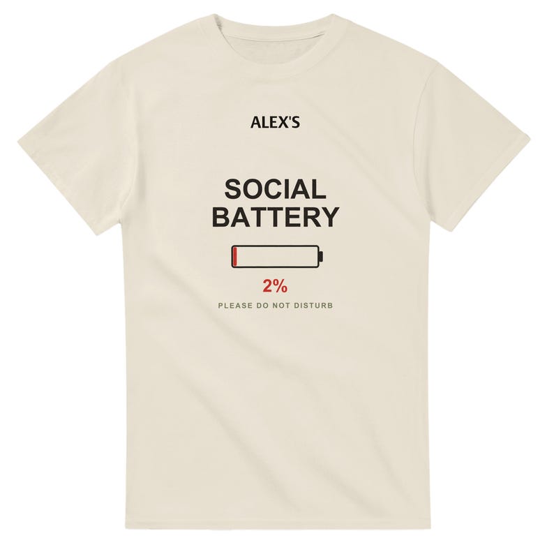 以下が含まれることがあります： クリーム色の半袖Tシャツ。前面中央に黒字で「ALEX'S SOCIAL BATTERY」、赤いバーが表示されたバッテリーアイコン、赤字で「2%」、黒字で「PLEASE DO NOT DISTURB」というテキストがプリントされています。