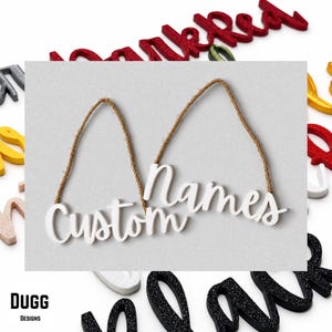 May include: Two white script-style word ornaments featuring the text Custom and Names, each attached to a brown twine hanging loop. The ornaments are displayed against a light gray background surrounded by various other glittery script words.