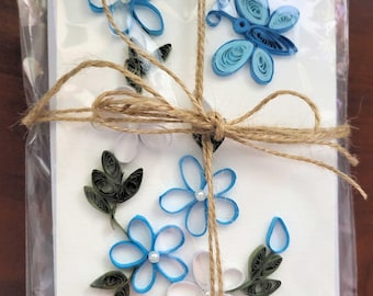 May include: A white greeting card decorated with blue and white paper quilling flowers, green leaves, and a blue butterfly, all featuring small white pearl centres. The card is wrapped in clear plastic and tied with a brown jute twine bow.