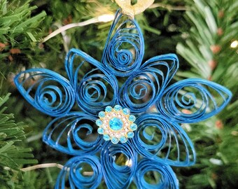 May include: A blue paper quilling ornament shaped like a star or flower, featuring intricate coiled paper loops and a central circular embellishment with small light blue rhinestones and a gold-tone rim, suspended by a thin gold ribbon loop.