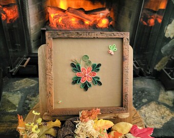 May include: A square wooden frame containing a paper quilling art piece featuring a central coral-coloured flower with dark green leaves and a small green butterfly accent. Three fabric pumpkins in brown, plaid, and yellow sit in front of the frame.