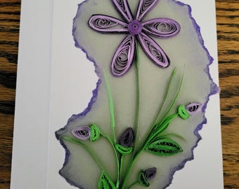 May include: A white greeting card featuring a paper quilling design of a purple flower with six petals, green stems, and leaves. The design is set against a translucent, irregular-shaped background with a purple-tinted, deckled edge.