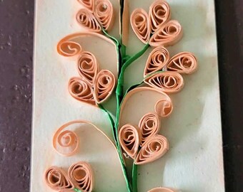 May include: A rectangular bookmark featuring a paper quilling design of a vertical green stem with peach-coloured coiled paper flowers. The floral elements are arranged in symmetrical clusters along the stem, creating a textured, three-dimensional effect on the light-coloured, smooth surface of the bookmark.